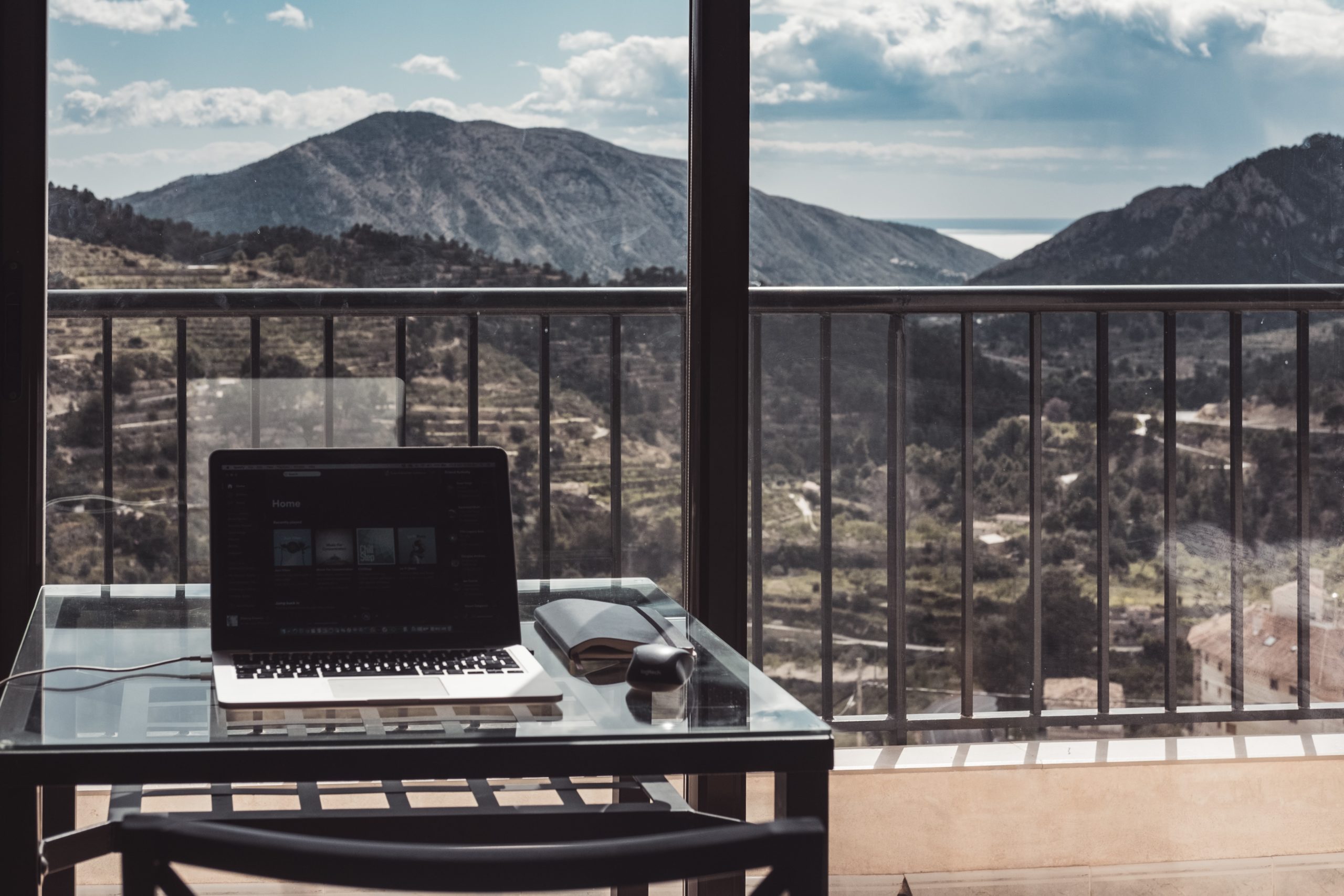 A Macbook rests on a desk, in front a window with a view of mountains and the sea.