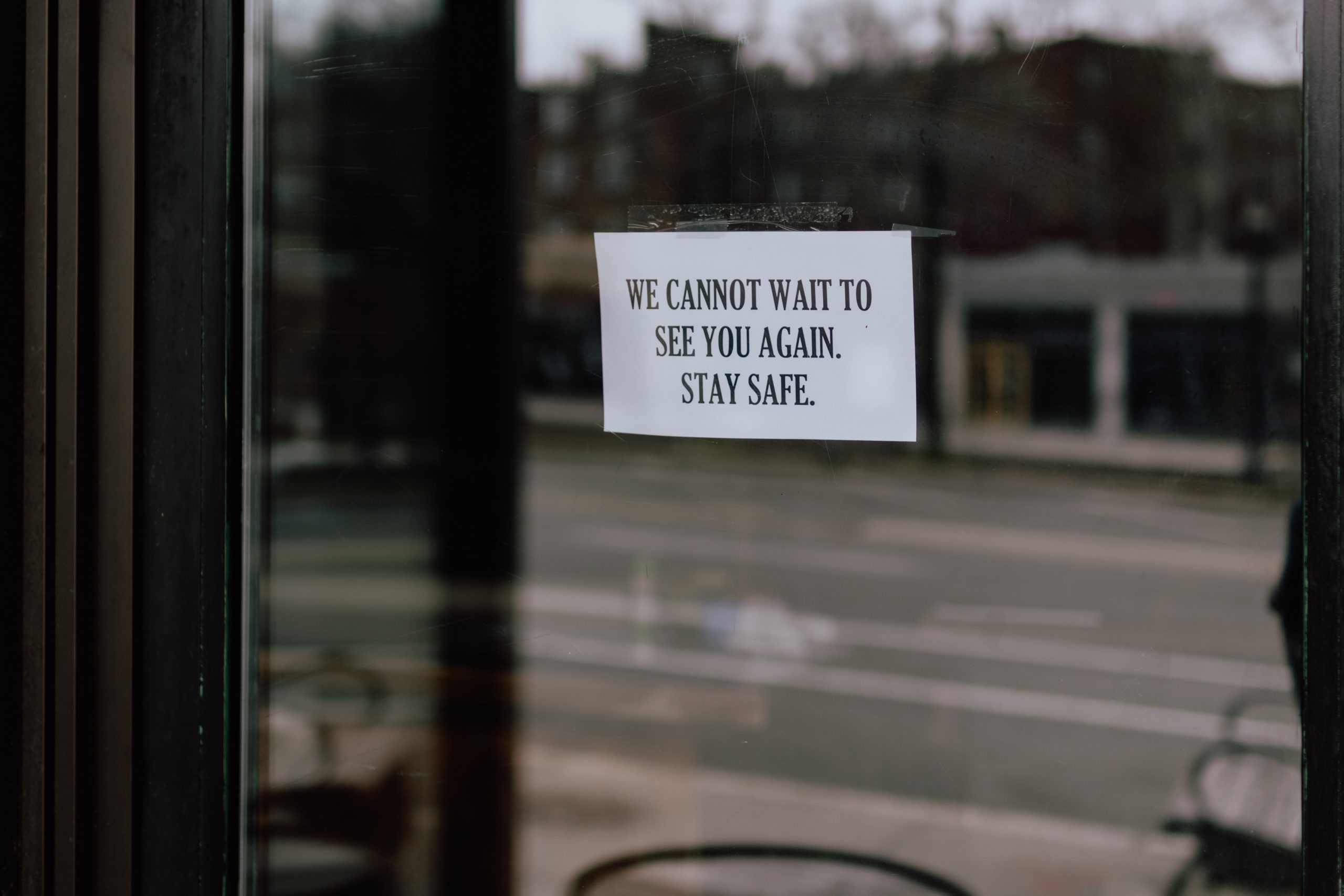 A business window displays a sign that says “We cannot wait to see you again. Stay safe