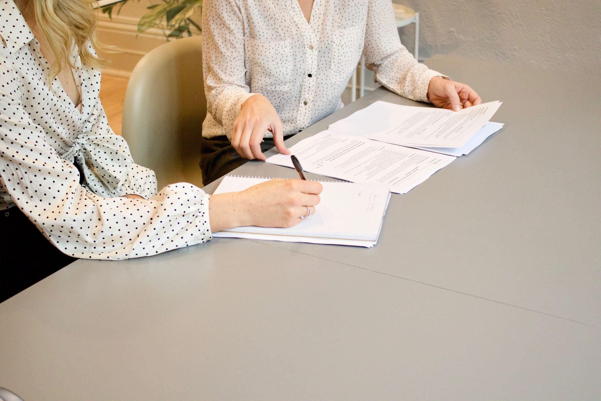Two women in business attire sit at a table reviewing and signing documents.