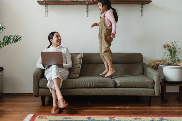 A woman working on a laptop on a couch at home while her daughter jumps next to her.