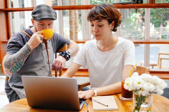 Young woman using a laptop while a man in a cap next to her sips coffee.