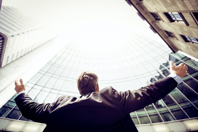 A man in a business jacket with outstretched arms looks up at a high rise office building.